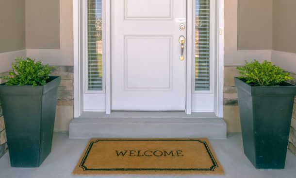 Alt="Front door with doormat plants and glass panel. White front door of a house with the word welcome printed on the brown doormat. Ornamental plants and glass panels are on each side of the door."