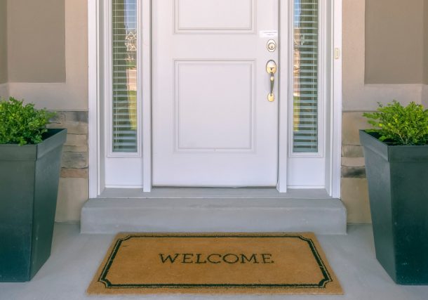 Alt="Front door with doormat plants and glass panel. White front door of a house with the word welcome printed on the brown doormat. Ornamental plants and glass panels are on each side of the door."