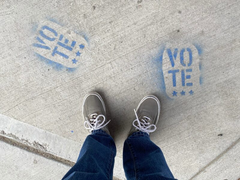 A young person's feet, and on the ground, sprayed in graffiti paint, the word 'Vote'