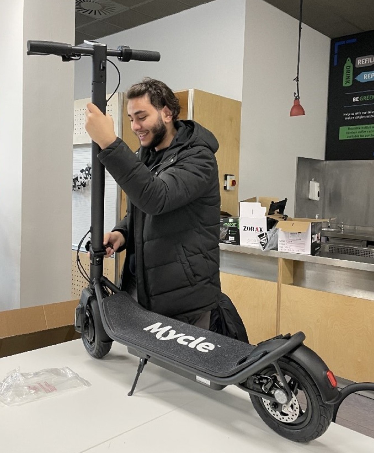 Male teenager in black coat holding onto the Mycle scooter he is building as it stands on a table