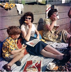 Two women and a child sat on a beach with a picnic with their backs against a wooden board.
