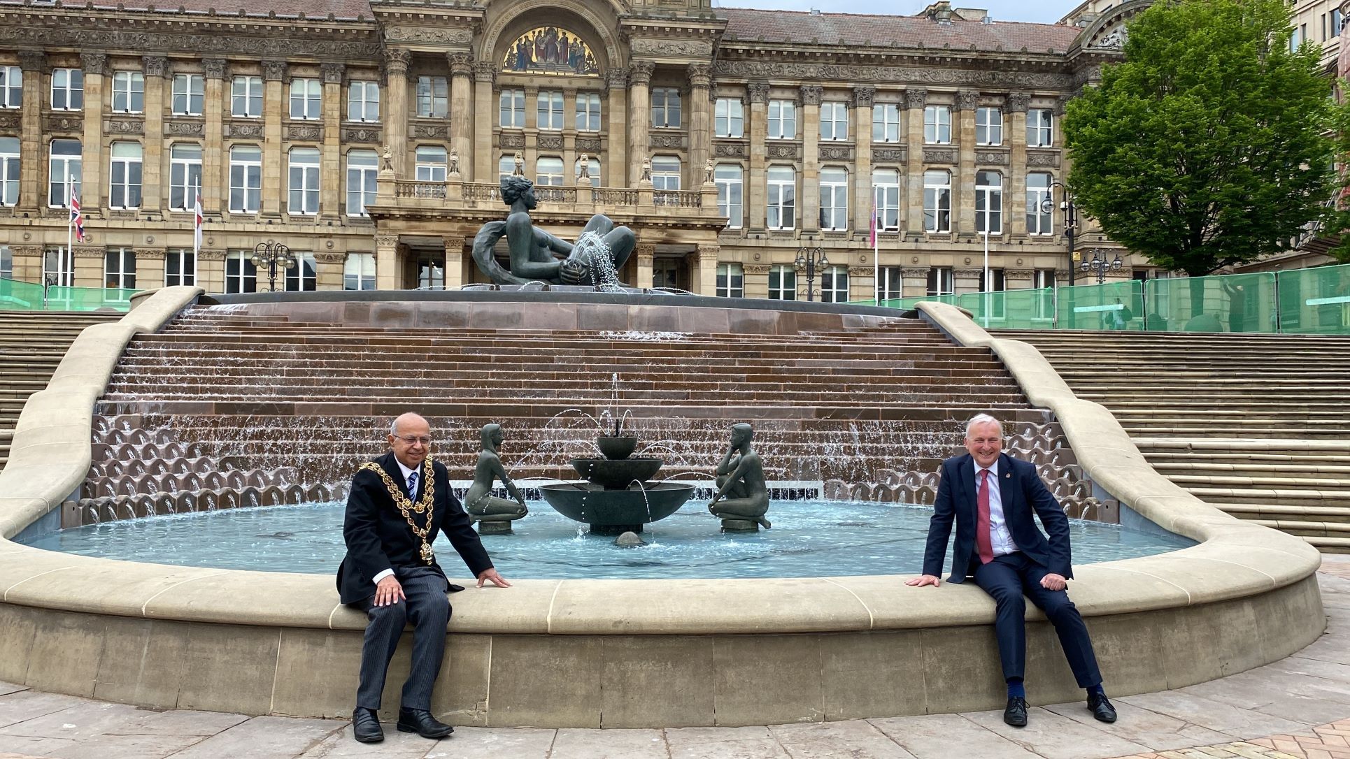Image of Lord Mayor Cllr Muhammad Afzal with Leader of the city council Cllr Ian Ward sat on the bottom ledge of The River fountain in Victoria Square - with the Council House in background