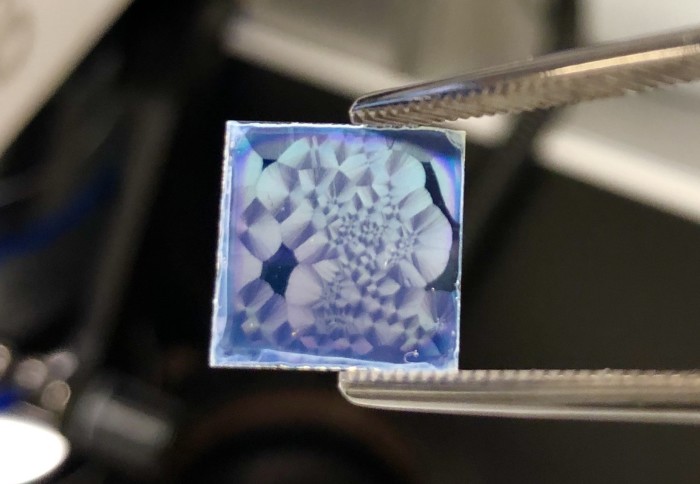 Tweezers holding a square of materials with seemingly random pattern of bluish reflective surfaces
