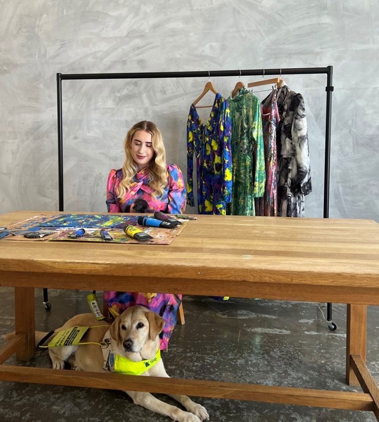Woman sitting at a table with a painting in front of her. A guide dog sits at her feet and behind is a rail of clothes with abstract prints.
