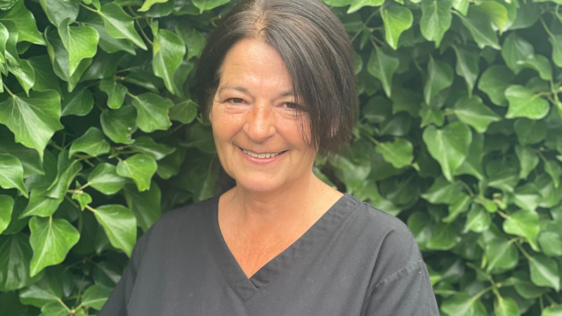 Lynn Sherlock stands smiling outside with greenery in the background, wearing a black tunic