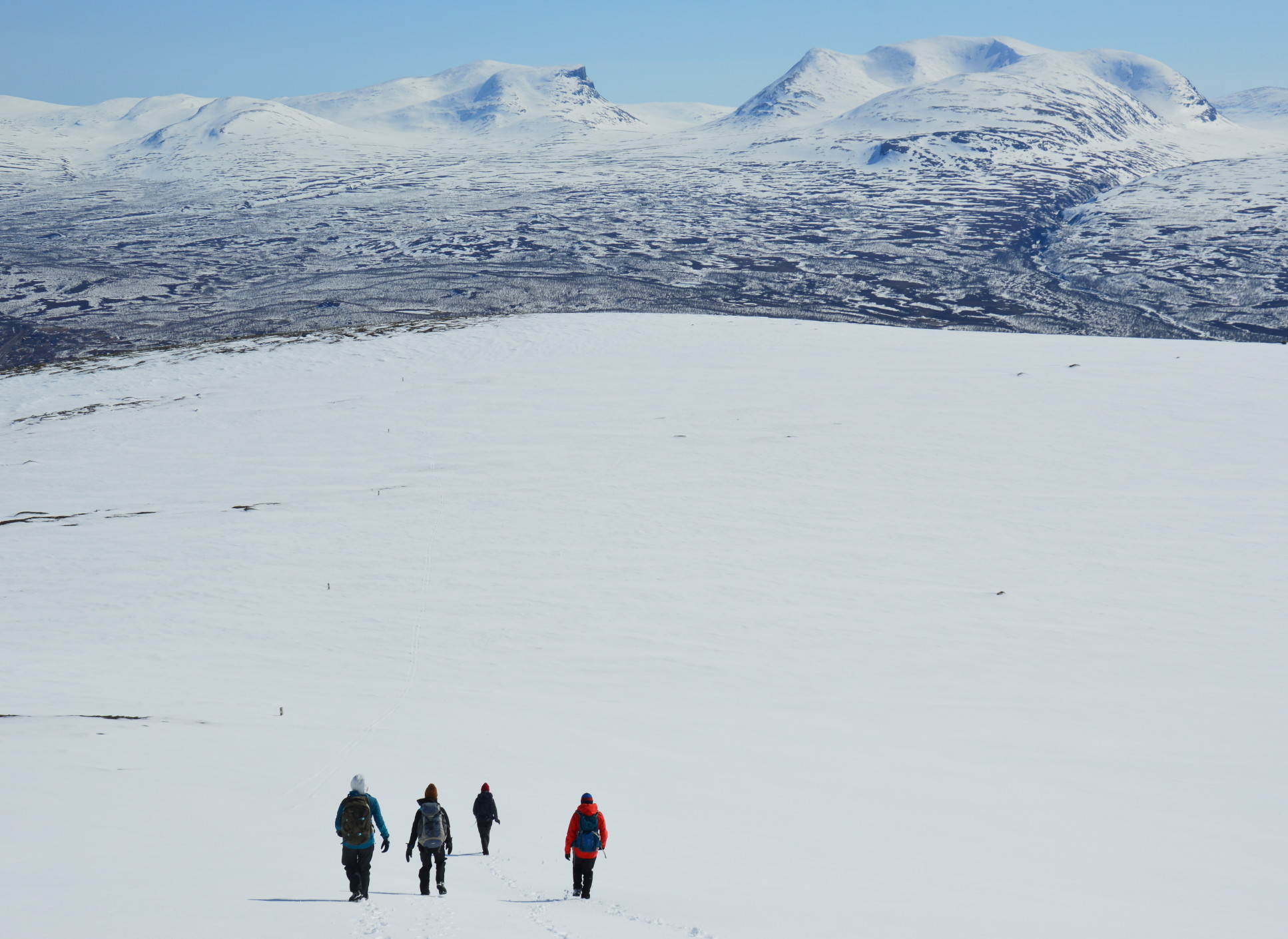 Four figures hiking across snow, with a wide tundra landscape in the distance
