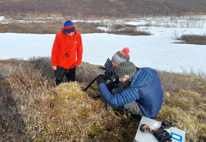 Man watching two people with cameras crouch over a mound of grass, with snow and a glacier in the background