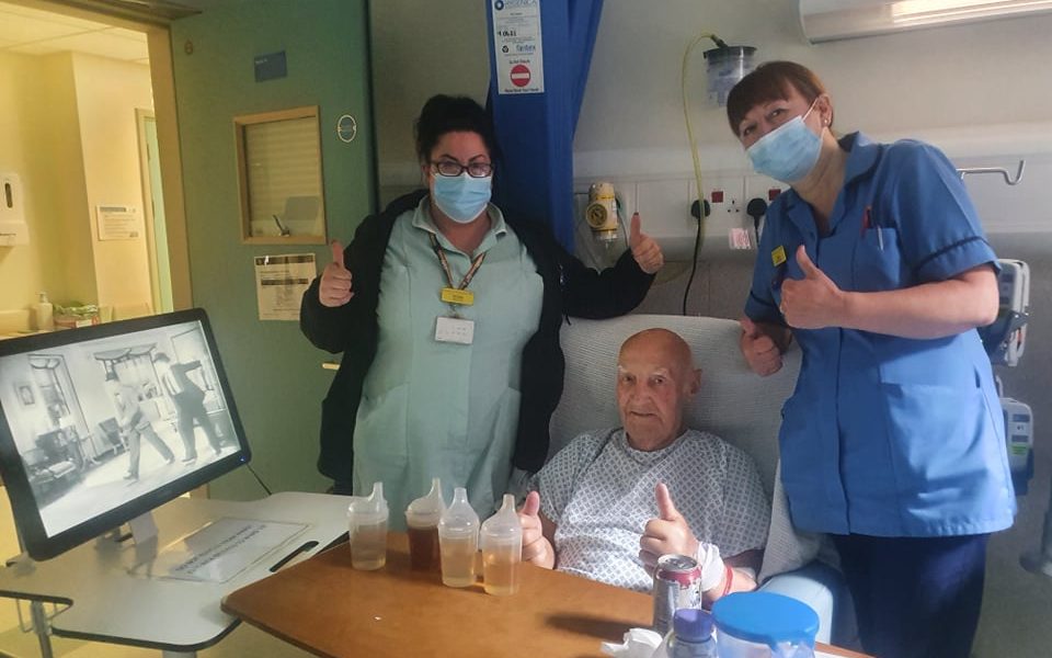 Two nursing staff stood next to a patient in bed with their thumbs up