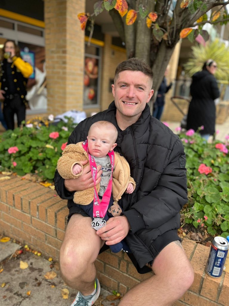 Man sat on a low wall in front of a flower bed with baby son on his lap. His son is wearing his brand new marathon medal around his neck.