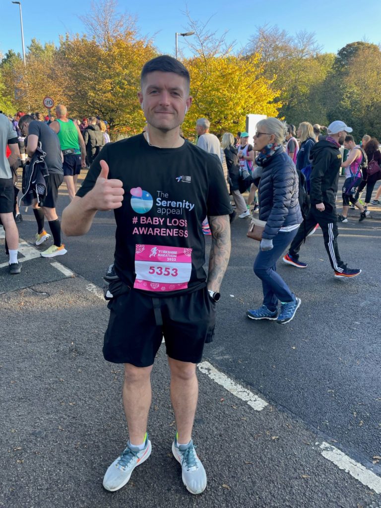 James is stood on the road giving a thumbs up sign to the camera. He is wearing a black t-shirt which has the text 'The Serenity Appeal #Baby Loss Awareness' on it. He is also wearing trainers, black running shorts and air pods. In the background there are crowds of people, including runners preparing for the marathon.