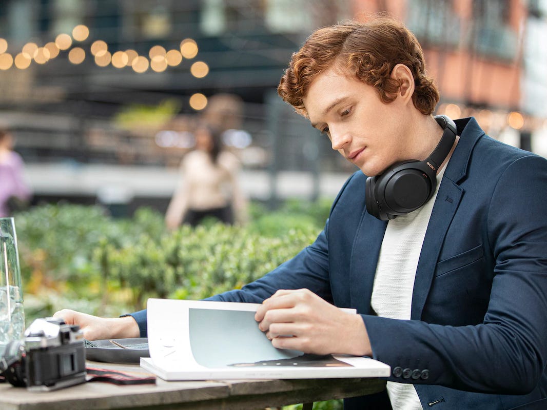 man reading at a cafe while wearing a pair of sony wh1000 xm4 headphones around his neck.