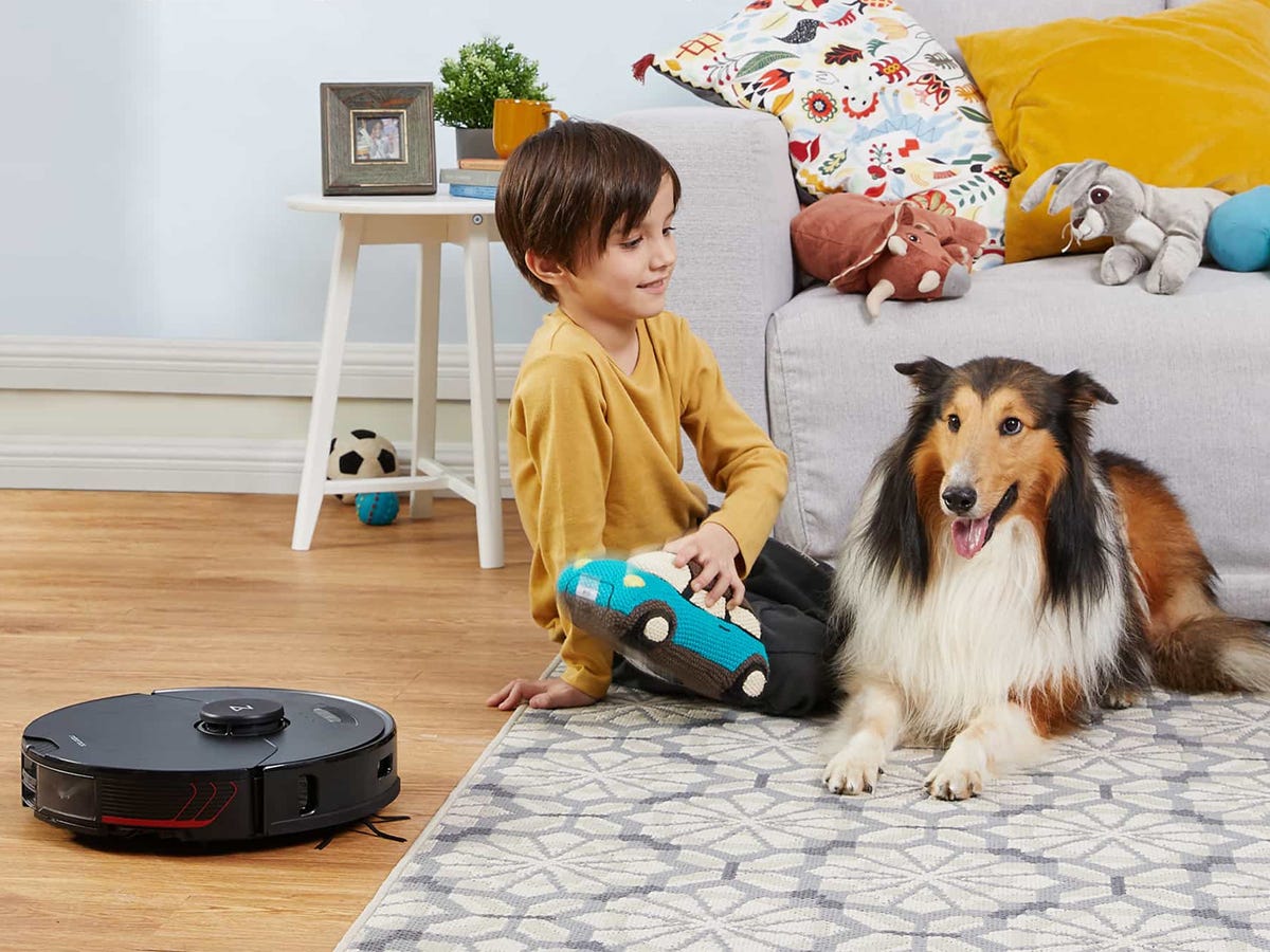 child sits and plays next to a collie dog in a living space with hardwood and an area rug as the Roborock S7 MaxV Ultra Robot Vacuum cleans the floor nearby