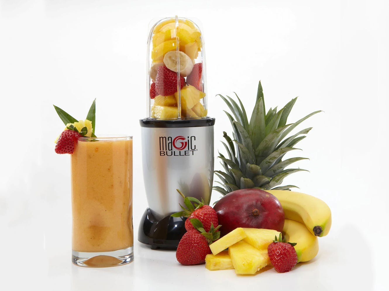 Magic Bullet blender with sliced fruit inside set next to a pile of whole fruit and a glass containing a smoothie.