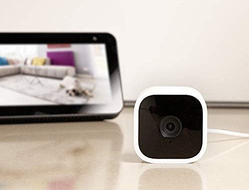 Blink Mini security camera set on a counter with its feed displayed on a tablet behind it.