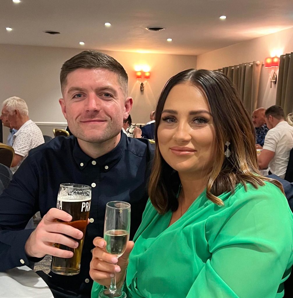 A man and a woman sat at a table together in a stylish venue, facing the camera, smiling and drinking alcohol
