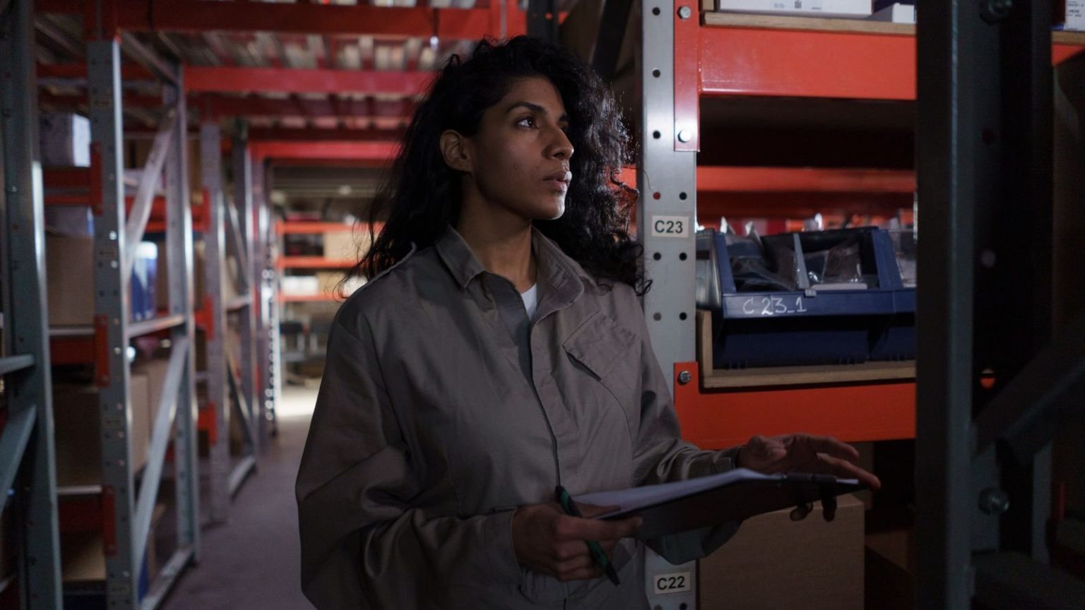 Rathbones finds 194 modern slavery reporting failures at FTSE 350 firms A person in a grey shirt holding a clipboard, standing in a large warehouse or storage facility with red shelving and metal structures.